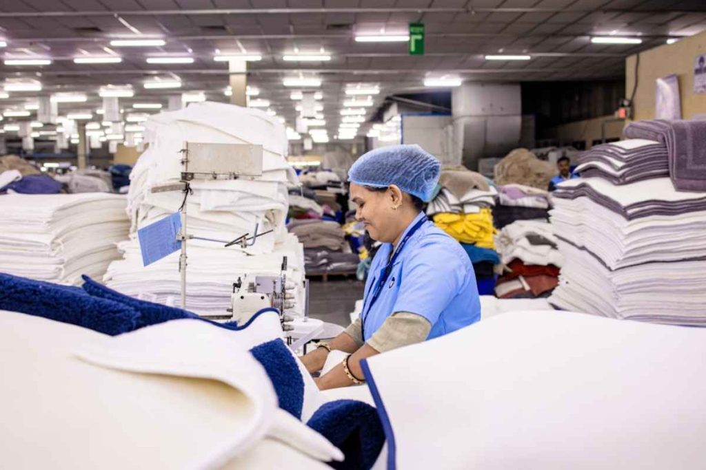A woman operates machinery in a factory surrounded by stacks of white towels, focusing on her task efficiently.